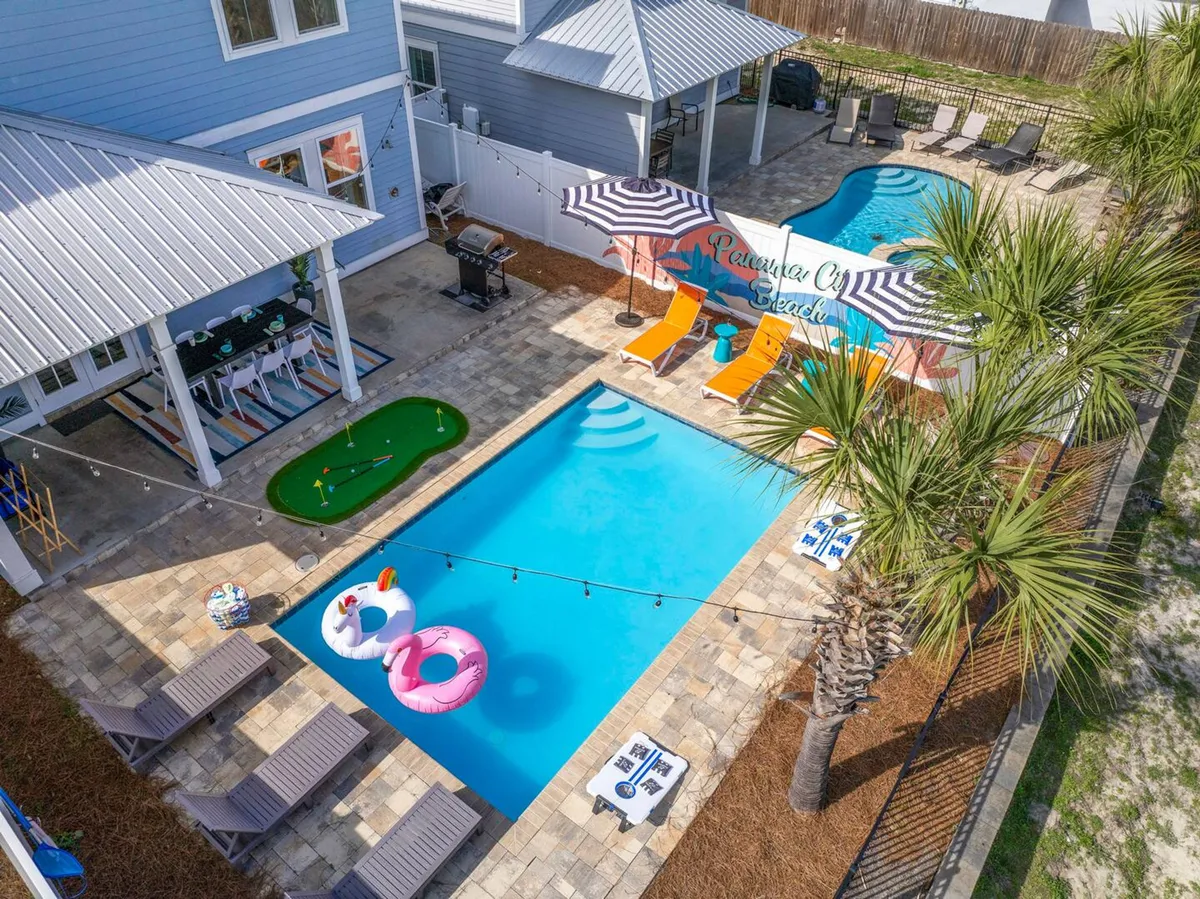 A lively backyard scene featuring a heated swimming pool with colorful flamingo and unicorn floats, an adjacent mini-putt green, a shaded outdoor dining area with bar stools under a covered patio, and a vibrant 'Panama City Beach' mural wall behind the loungers. The area is well-lit, festive, and designed for both relaxation and active fun.