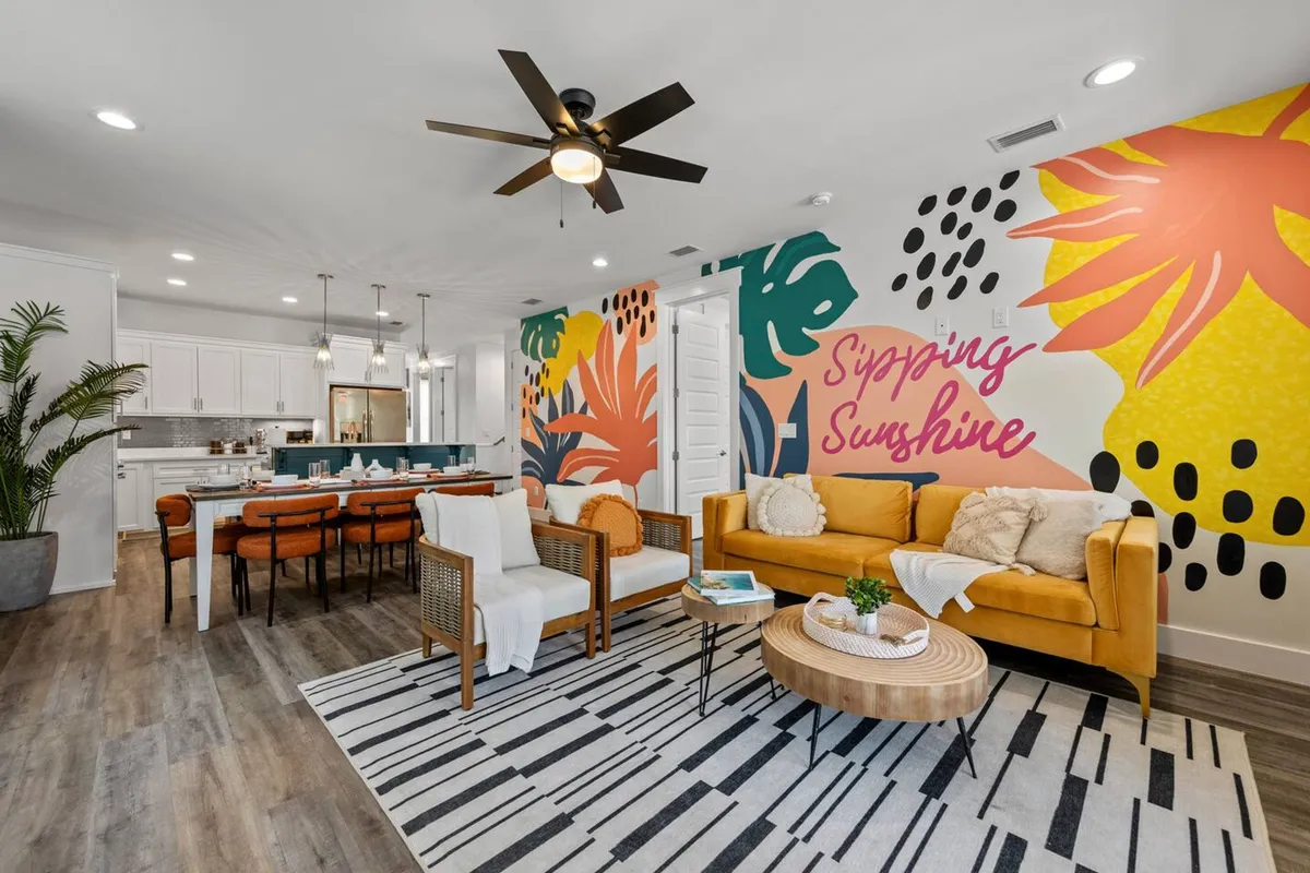 This image features a bold, colorful wall mural with tropical leaves and 'Sipping Sunshine' text as the backdrop to a mustard yellow sofa adorned with cozy pillows. A modern dining table with leather chairs sits nearby, and a ceiling fan with a light fixture completes the space. The open-plan layout clearly connects the living and dining areas, illuminated by natural light, and hints at the adjacent kitchen.