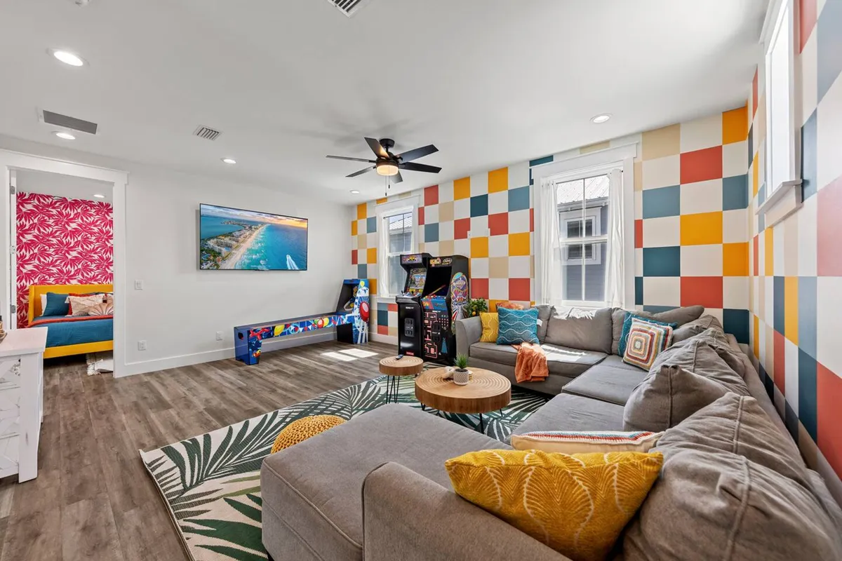 A playful, energetic room with a colorful checkerboard accent wall, a large gray sectional sofa with patterned pillows, and arcade game machines, including pinball and shuffleboard tables. The space is designed for both relaxation and lively entertainment, providing ample seating for group gatherings.