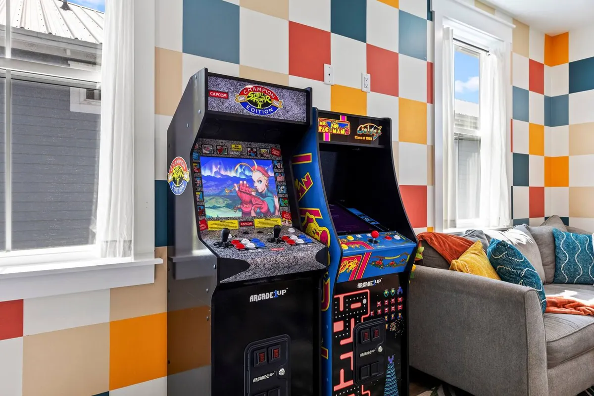A bright, playful indoor space featuring classic arcade game machines (Street Fighter and Ms. Pac-Man), a comfortable gray couch with colorful pillows, and large windows letting in natural light. The ambiance is energetic and nostalgic, inviting guests to enjoy indoor entertainment.
