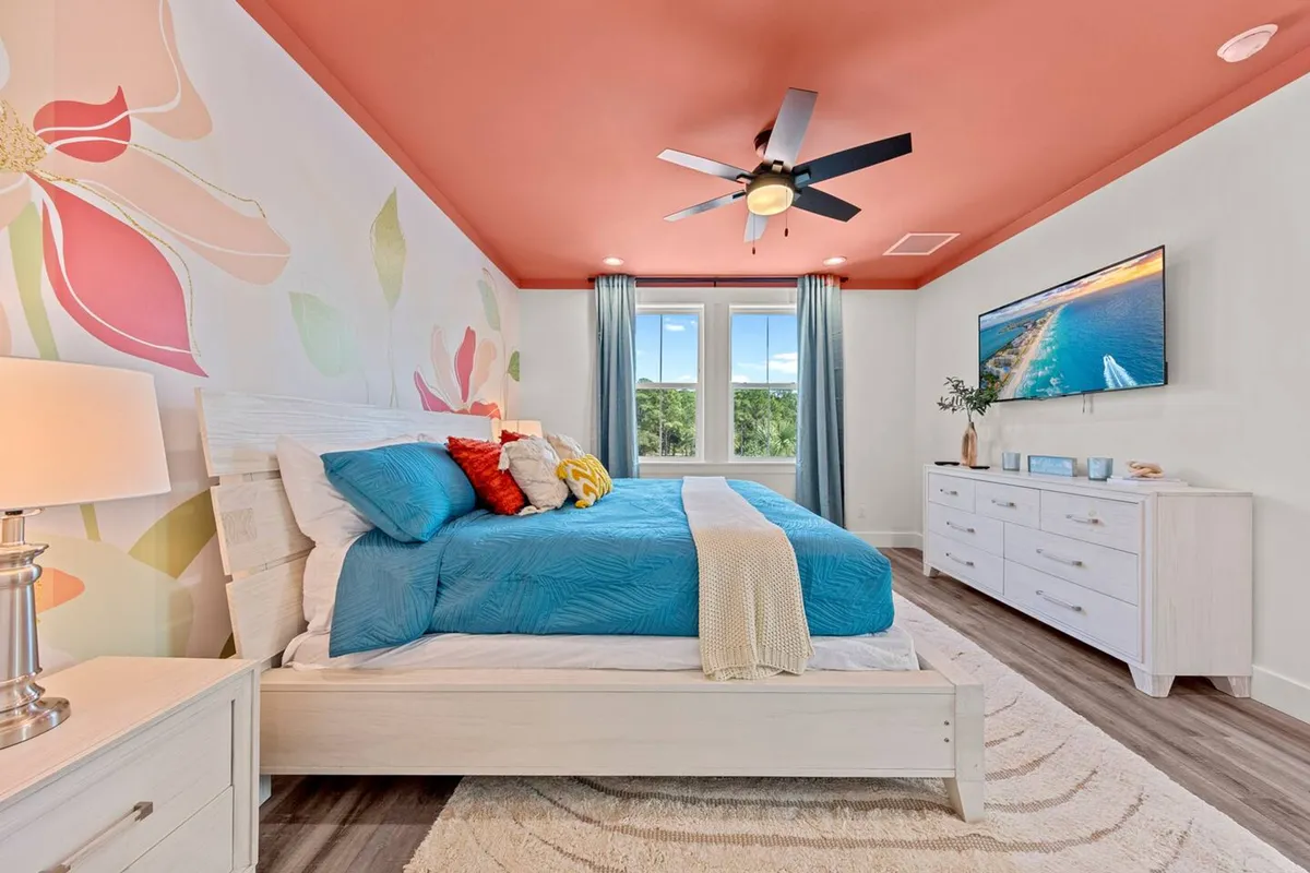 A cheerful bedroom featuring a king-sized bed with blue bedding, colorful pillows, and a floral accent wall. The room includes a modern ceiling fan, a white dresser with a wall-mounted TV, and ample natural light from a window, conveying a sense of comfort and relaxation.