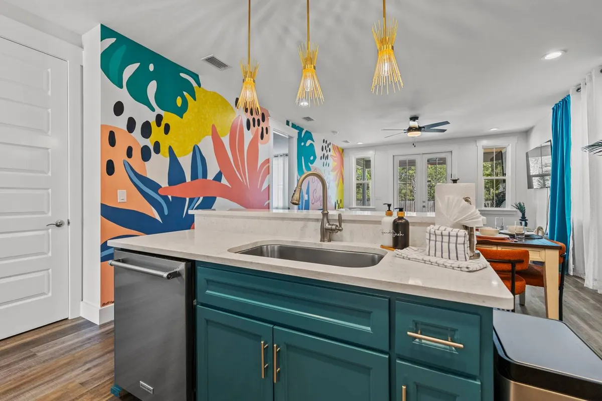 A cheerful, inviting space featuring a colorful tropical mural wall, a teal kitchen island with a sink and brass pendant lights, and a dining area with blue curtains. The design encourages social interaction, with the kitchen and dining area flowing openly into the living space.