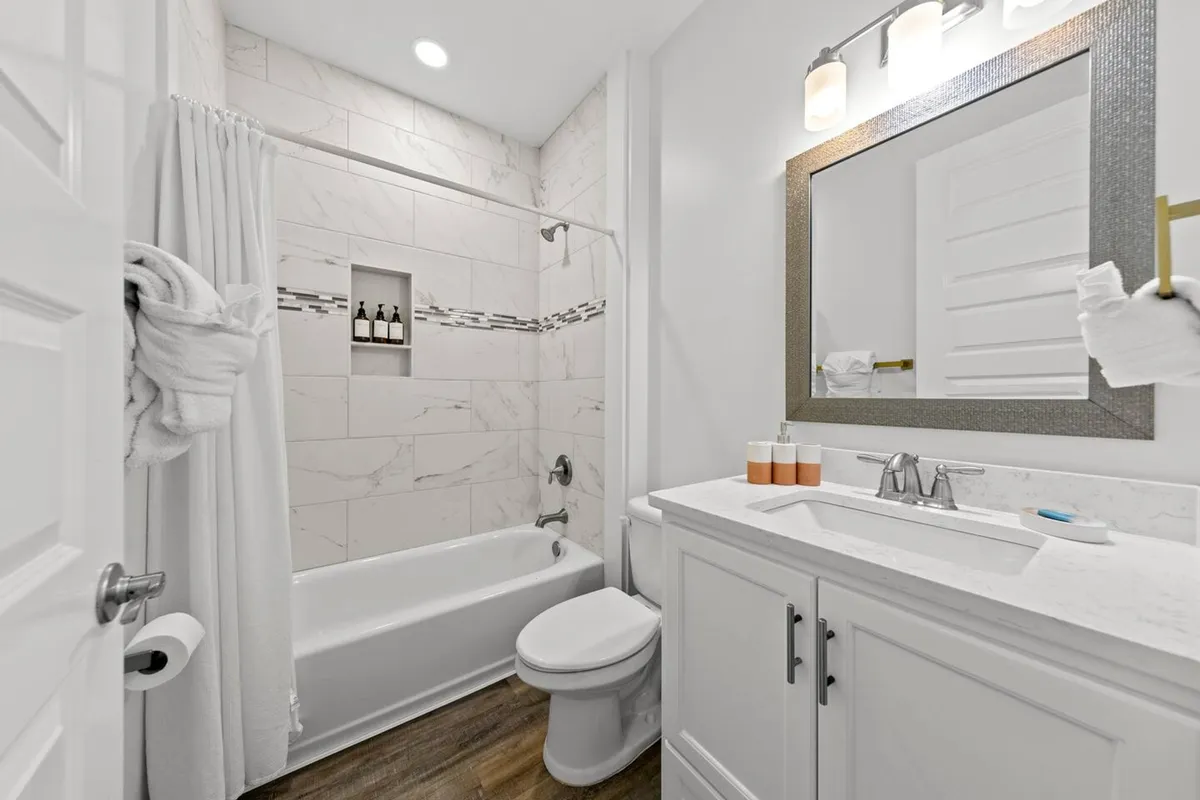 A fresh, modern bathroom with a marble-tiled bathtub/shower combo, a large framed mirror above a white vanity with a marble countertop, and neatly folded white towels and toiletries. The space is bright, clean, and inviting.