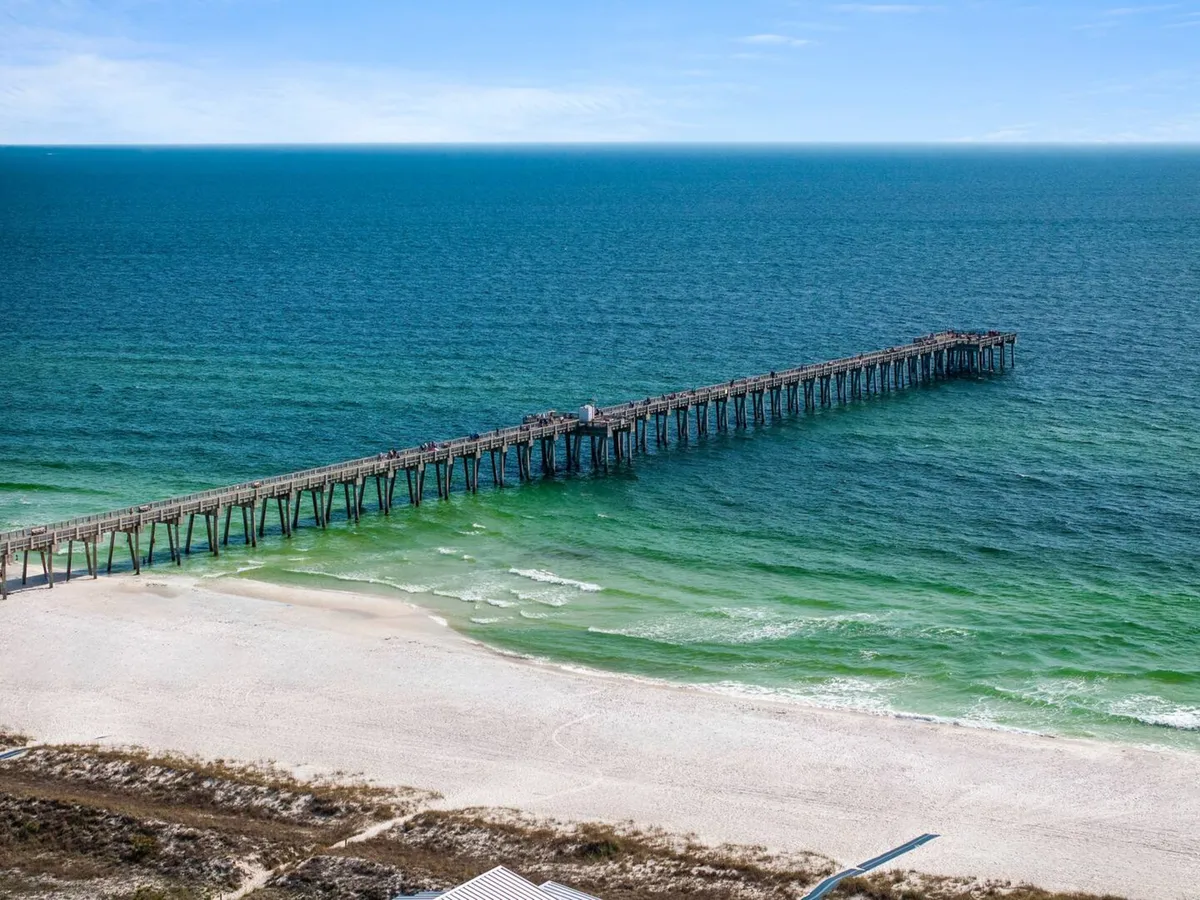 A long wooden pier stretches out into the vibrant blue-green ocean, flanked by pristine white sandy beaches. The scene invites guests to enjoy classic Panama City Beach activities, such as fishing, strolling, or gathering by the water for sun and fun.