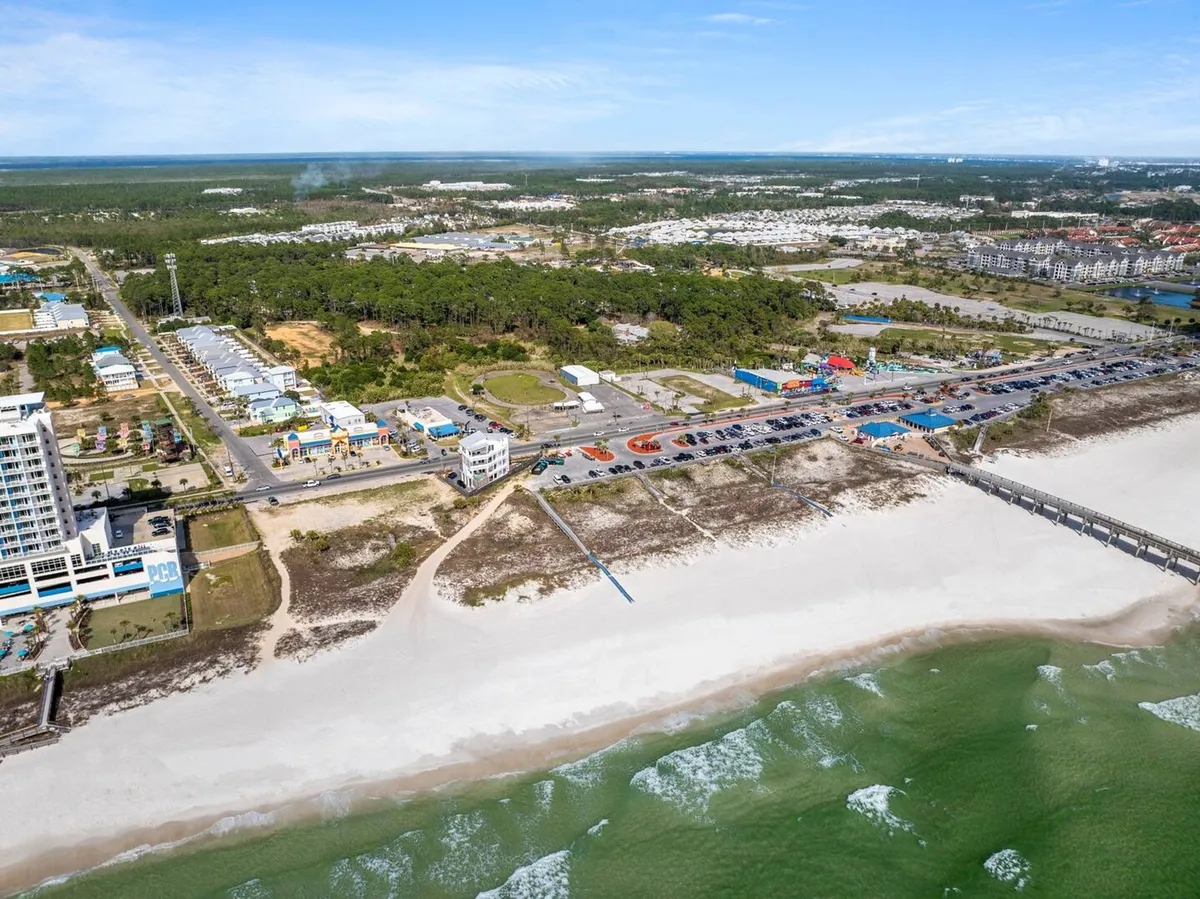 A lively beach scene featuring a wide stretch of sandy shoreline, rolling ocean waves, a pier stretching into the water, and rows of beachfront properties. Beachgoers are seen walking, sunbathing, and enjoying the vibrant local beach atmosphere.